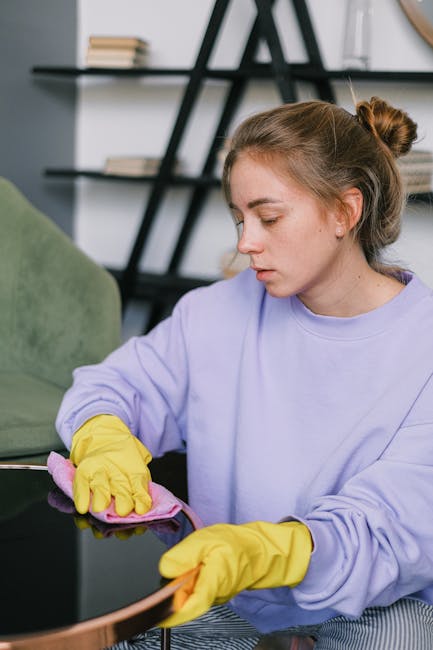 A young woman with light brown hair tied in a bun, wearing a lavender sweatshirt and yellow rubber gloves, is vacuuming a black, shiny glass desktop surface in a modern living room. The room features a green upholstered chair, a black shelving unit with books and decorative items in the background, and natural light illuminating the space. This scene exemplifies professional domestic cleaning, focusing on surface cleaning and dust removal, aligned with the services offered by Kensington Carpet Cleaners as highlighted on the page about affordable end of tenancy cleaning costs in Kensington W8.