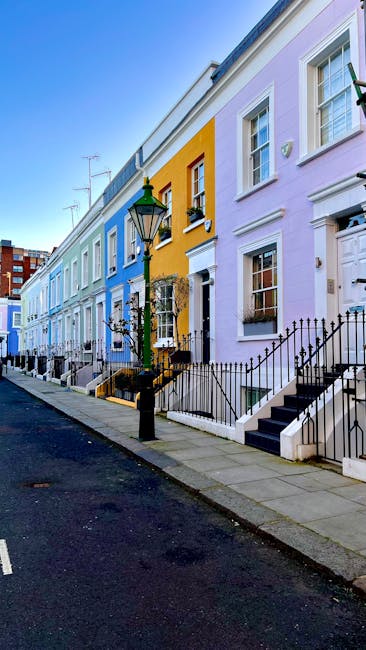 A row of colorful terraced houses in Kensington W8 featuring pastel-colored facades in shades of pink, lavender, yellow, and blue. The houses have white-framed windows with decorative window sills, and black wrought-iron railings along small front steps leading to front doors. The street is paved with a smooth asphalt surface, with a traditional lamppost near the curb. The scene is illuminated by natural daylight, highlighting the clean and well-maintained appearance of the building exteriors, which are part of a residential area. While no interior surfaces are visible, the exterior setting suggests a charming, tidy environment consistent with professional cleaning standards offered by Kensington Carpet Cleaners, especially relevant for end of tenancy cleaning in Kensington W8.