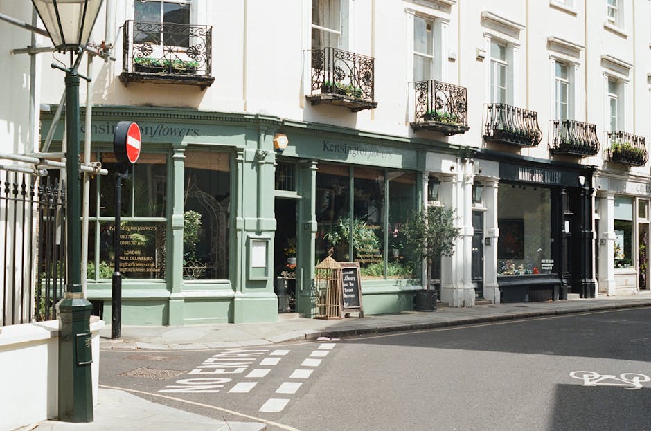 The image depicts the exterior of a charming, multi-story commercial building on a street corner, featuring a green-painted storefront with large glass windows displaying potted plants and floral arrangements. The upper floors have white facades with black wrought iron balconies adorned with flower boxes. The sidewalk in front is clean and well-maintained, with a small blackboard sign placed outside the shop, and a no-entry traffic sign visible at the corner. The street surface appears smooth, with a designated bike lane nearby, and soft daylight illuminates the building, highlighting its elegant architectural details. This setting exemplifies a well-maintained commercial space in Kensington, emphasizing cleanliness and upkeep, as promoted by Kensington Carpet Cleaners for business premises.