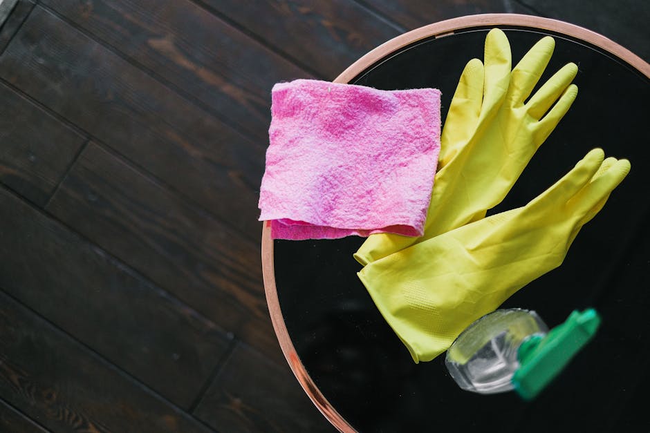 A person wearing yellow rubber gloves is placing a litup sign that reads 'CLEANING HOME' onto a wooden surface in a modern residential or commercial space, with visible wooden flooring, glass windows,