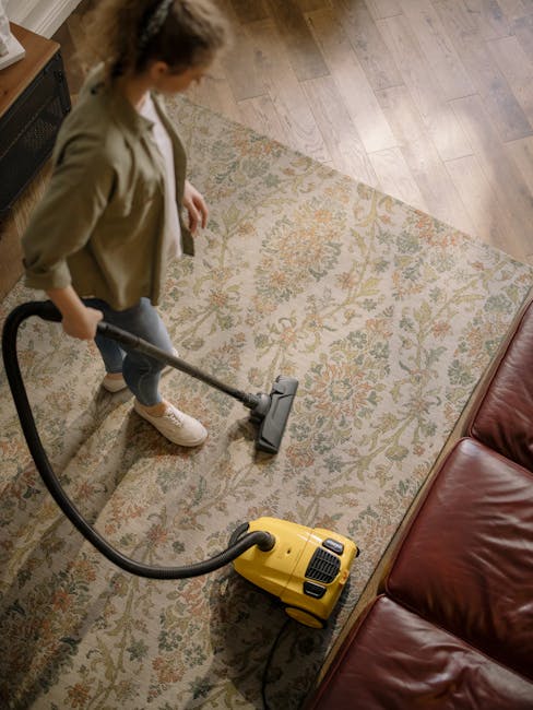 A woman is engaged in surface cleaning using a yellow vacuum cleaner with a black hose, vacuuming a large, ornate, cream-colored rug with floral patterns in a living room. The scene features a wooden floor surrounding the rug and a red leather sofa positioned on the right. The room is well-lit with natural light, highlighting the cleanliness and condition of the rug and surrounding surfaces. This image illustrates domestic cleaning practices, emphasizing thorough carpet and surface maintenance, consistent with the services offered by Kensington Carpet Cleaners highlighted in the article about hidden carpet cleaning charges to avoid in Kensington.