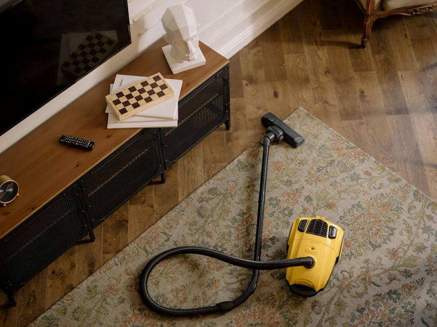 A yellow vacuum cleaner with a long hose attachment is positioned on a decorative area rug in a room with wooden flooring. To the left, a black metal and wood media console holds a stack of papers, a chessboard, a remote control, and a white bust sculpture. The room is brightly lit, with the wooden floor showing a polished, dust-free surface, and the area rug displaying a detailed floral pattern. This scene highlights routine domestic cleaning, with an emphasis on surface cleaning and maintaining hygiene inside a living space, as part of professional cleaning services offered by Kensington Carpet Cleaners.