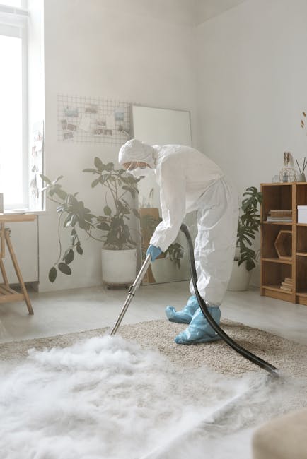A professional cleaning technician dressed in a white protective suit, hood, mask, and orange gloves using a portable vacuum cleaner to conduct surface cleaning and sanitisation in a modern indoor set