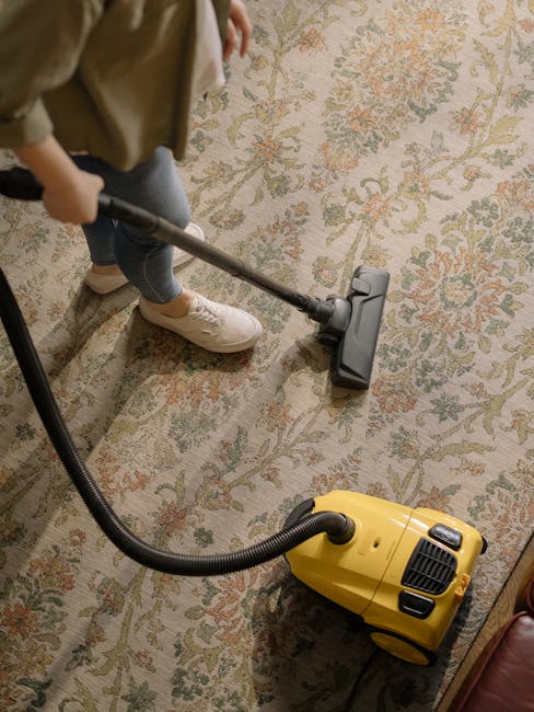 A person using a vacuum cleaner with a yellow body and black hose attachment on a traditional patterned carpet in a residential setting. The carpet features floral motifs in muted shades of beige, green, and orange. The individual, dressed in casual clothing including beige shoes and jeans, is standing on the carpet while performing surface cleaning. The room appears well-lit, highlighting the clean and maintained condition of the carpet, with the vacuum actively engaged in deep cleaning to remove dust and dirt. This scene exemplifies professional domestic cleaning services provided by Kensington Carpet Cleaners, aligning with their focus on hygiene and surface maintenance for carpeted areas.