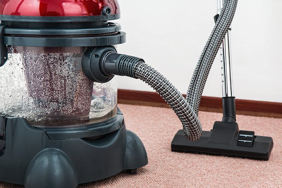 A person using a vacuum cleaner with a yellow body and black hose attachment on a traditional patterned carpet in a residential setting. The carpet features floral motifs in muted shades of beige, green, and orange. The individual, dressed in casual clothing including beige shoes and jeans, is standing on the carpet while performing surface cleaning. The room appears well-lit, highlighting the clean and maintained condition of the carpet, with the vacuum actively engaged in deep cleaning to remove dust and dirt. This scene exemplifies professional domestic cleaning services provided by Kensington Carpet Cleaners, aligning with their focus on hygiene and surface maintenance for carpeted areas.