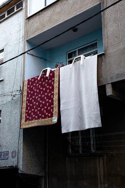 External view of an apartment building showing a clothesline strung across the balcony, with a patterned red and gold rug and a plain white towel hanging to dry. The building’s facade features textured concrete and rectangular windows, one of which is open with a white curtain visible inside. The scene is illuminated by natural daylight, highlighting the weathered surfaces and the drying laundry, exemplifying typical residential exterior spaces. This image relates to surface cleaning and maintenance as part of routine domestic hygiene, consistent with Kensington Carpet Cleaners' services discussed in the Kensington High Street Rug Cleaning Guide for Local Homes.