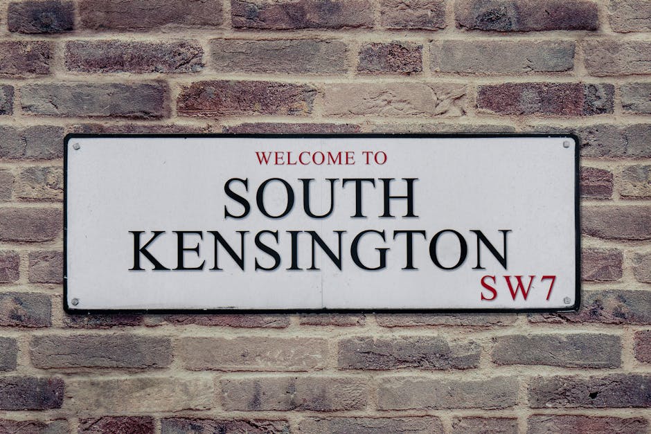 A rectangular metal street sign mounted on a red brick wall, displaying the words 'Welcome to South Kensington' in black uppercase letters, with 'SW7' in red at the bottom right corner. The sign is positioned centrally against the textured bricks, which vary in shade from light to dark red. The lighting is natural, illuminating the sign clearly, and the overall scene conveys a clean and well-maintained urban environment, reflective of the area serviced by Kensington Carpet Cleaners for surface cleaning, deep cleaning, and sanitisation in local homes.