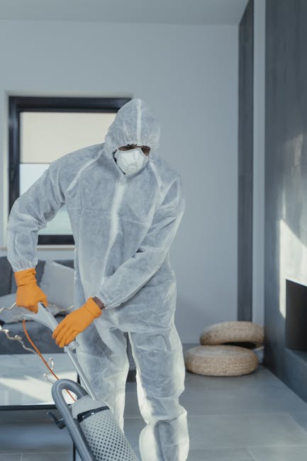A professional cleaner dressed in a white protective suit, face mask, and orange gloves is operating a professional carpet cleaning machine in a modern, well-lit living room. The room features large w