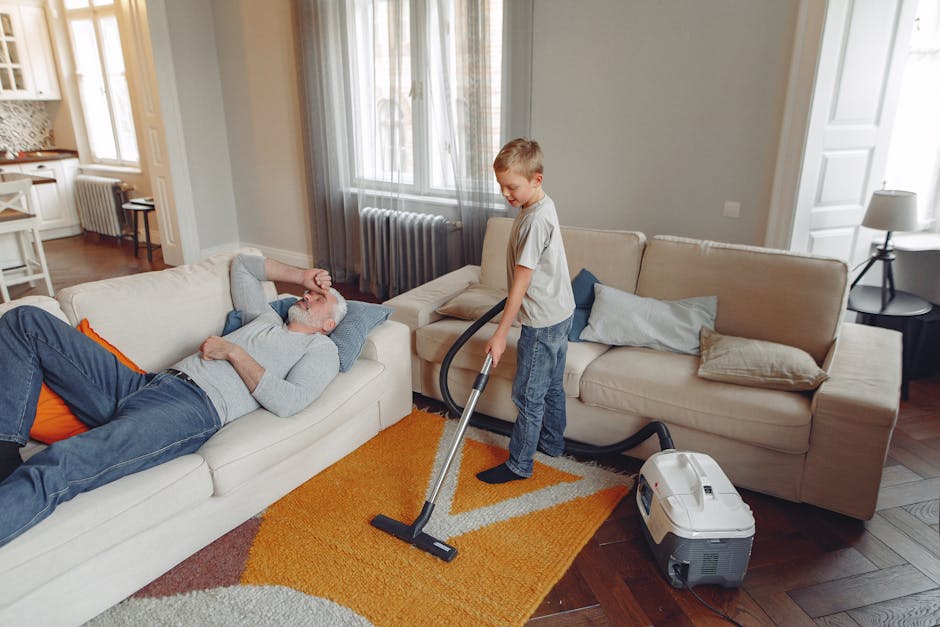 A young boy vacuuming a beige sofa in a living room during a cleaning session, while an older man with grey hair and beard is lying on the sofa, resting with his eyes closed. The living room features hardwood flooring with an orange and beige area rug, two large windows with sheer curtains allowing natural light to fill the space, and light-colored walls. Visible furniture includes a beige sofa with multiple cushions, a side table with a lamp, and a radiator beneath the windows. The boy uses a handheld vacuum cleaner connected to a portable vacuum unit placed on the floor nearby, demonstrating a routine surface cleaning process. The room appears clean and tidy, emphasizing domestic cleaning and maintenance in a residential setting, with natural daylight highlighting the hygienic condition of the space as part of a deep cleaning service by Kensington Carpet Cleaners.