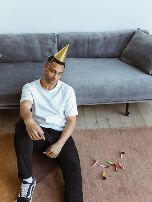 A young man with light skin, wearing a white t-shirt, black pants, and a gold party hat, sits on the floor beside a gray fabric sofa in a living room. The sofa has a smooth, slightly shiny surface and is positioned against a plain white wall. The man appears to be looking down, with a neutral expression. On the floor, in front of him, are some scattered discarded party blowers and confetti. The room is illuminated by natural light, highlighting the clean, tidy appearance of the space. The wooden floor beneath the carpet is visible, and the overall scene suggests a setting after a celebration or party, with attention to cleaning and hygiene by Kensington Carpet Cleaners, reflecting the focus on surface cleaning and deep cleaning relevant to upholstery after parties near Royal Albert Hall.