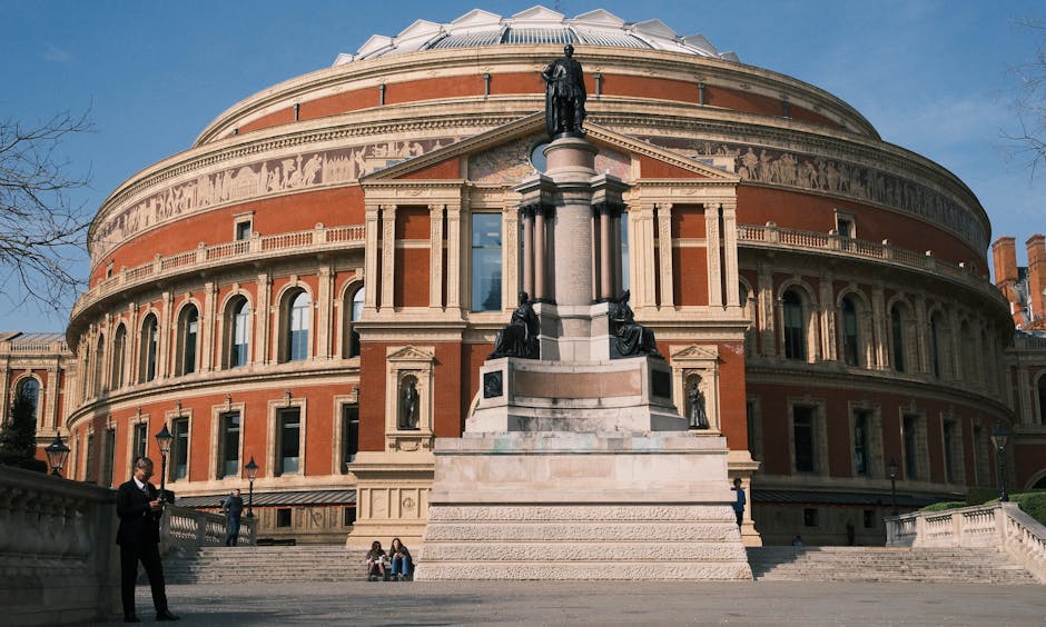 Exterior view of the Royal Albert Hall with its distinctive circular brick façade and ornamental detailing, taken during daytime with clear blue sky. In the foreground, a statue with multiple figures stands atop a pedestal, surrounded by steps leading to the hall entrance. Several visitors, including a man in a suit reading a tablet and a few individuals seated on the steps, are visible. The area is illuminated by natural light, highlighting the building's architectural features and the clean, well-maintained stone surfaces. This image showcases a historic cultural venue near Kensington, relevant to domestic cleaning discussions focused on surface maintenance and cleaning after events at such locations.